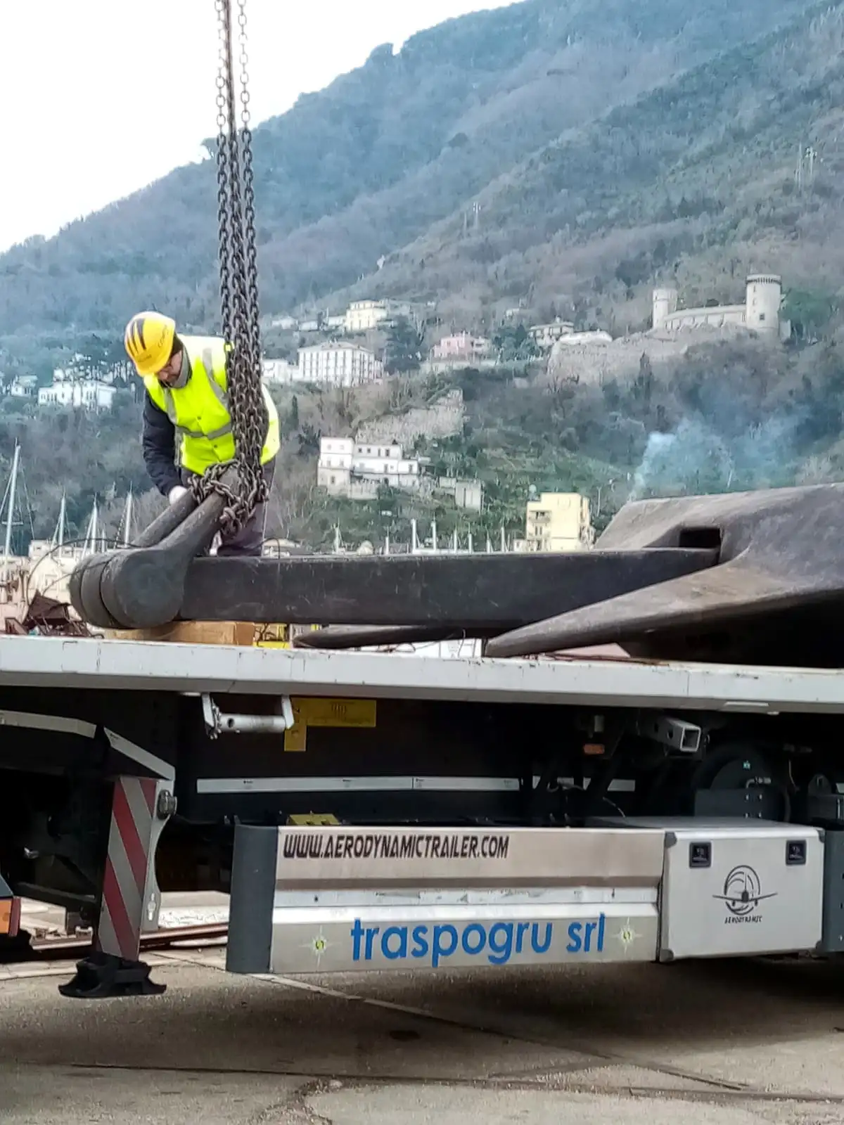 Operatore con casco imbraca un’ancora navale sul pianale Traspogru, con il castello sulla collina sullo sfondo