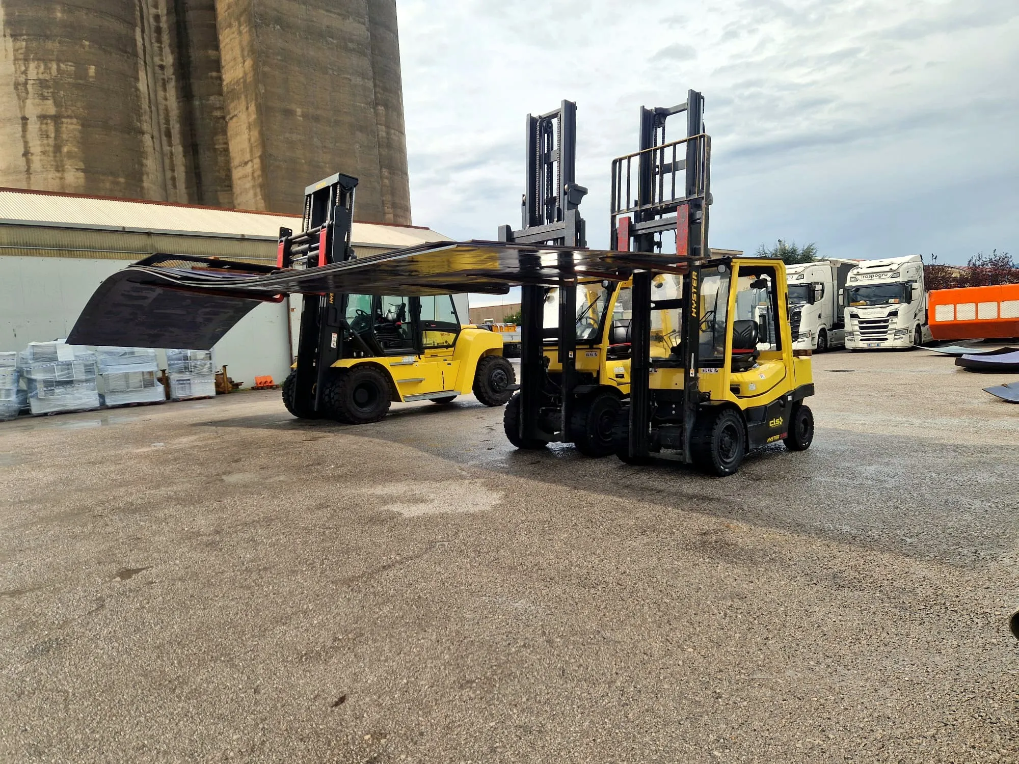 Due carrelli elevatori gialli sollevano insieme una grande lamiera nel piazzale, con altri mezzi sullo sfondo