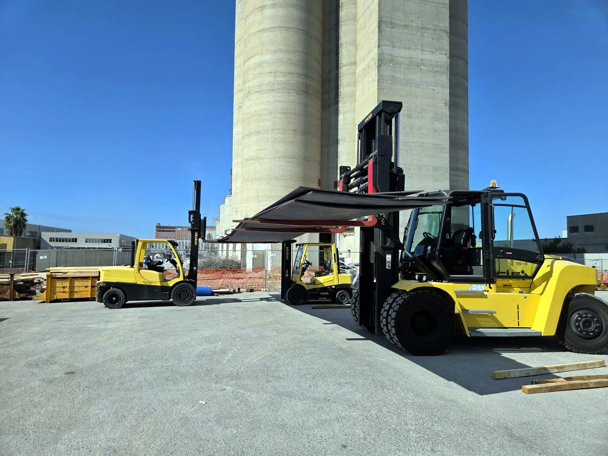 Due carrelli elevatori gialli sollevano insieme una grande lamiera nel piazzale
