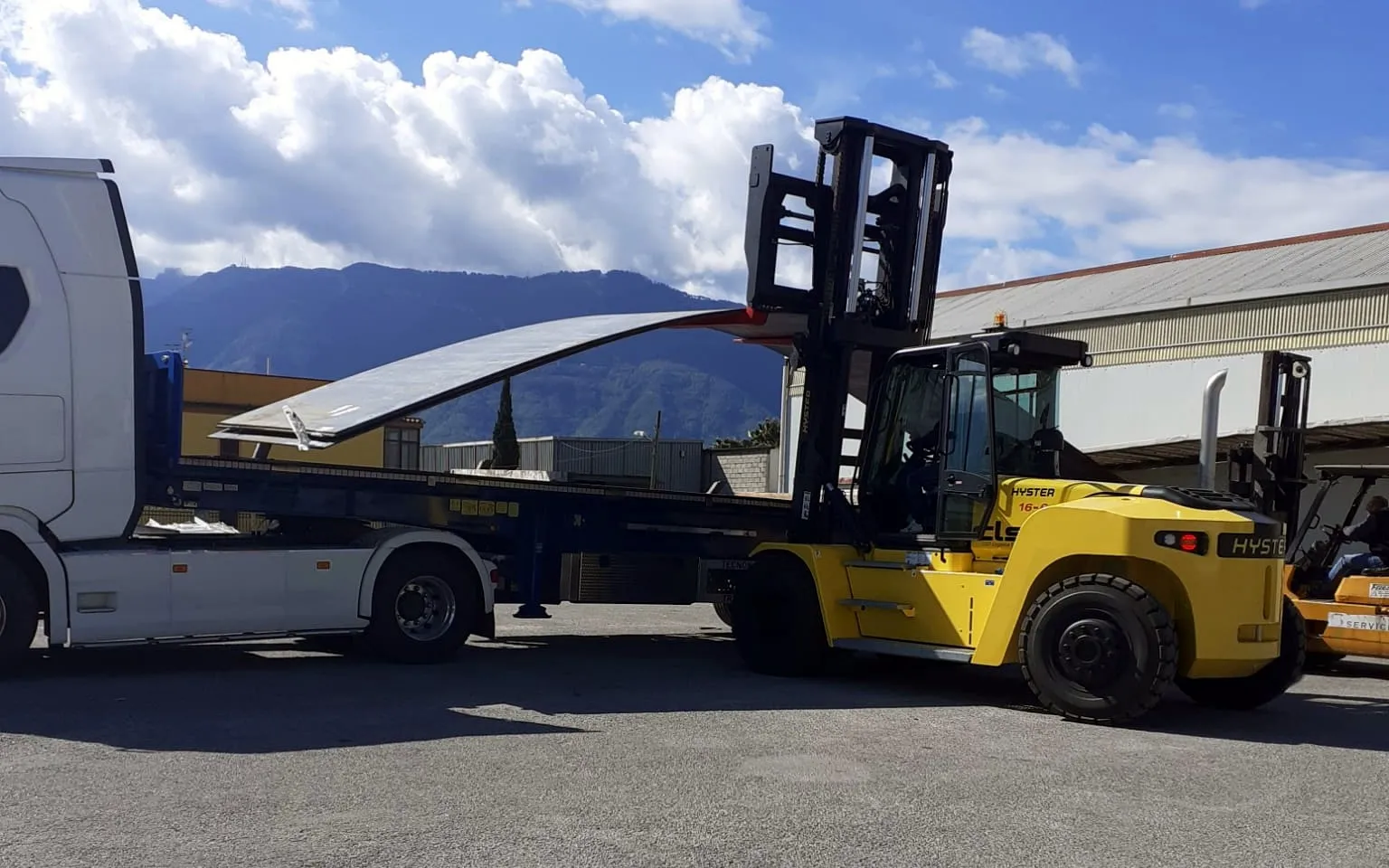 Carrello elevatore giallo da 16 tonnellate carica una grande lamiera su un semirimorchio, con il monte sullo sfondo